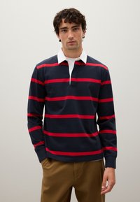 Uomo con capelli ricci scuri, che indossa una maglia da rugby a maniche lunghe di colore blu navy con ampie righe rosse e colletto bianco, abbinata a pantaloni marroni, sfondo neutro.