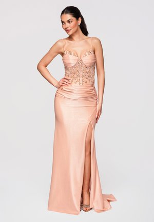 Femme en robe longue en satin pêche avec des perles détaillées sur le corsage et une fente haute à la cuisse, posant avec une main sur la hanche.