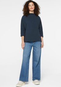 Navyblaues, strukturiertes Oberteil mit Dreiviertelärmeln, kombiniert mit weiten blauen Jeans und weißen Sneakers. Einfaches, zeitgenössisches Design und Passform.