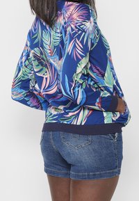 Femme portant une veste bleue à imprimé tropical avec des poches et un short en jean, les mains dans les poches de la veste, tournée dos à la caméra.