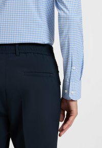 Camicia a quadri blu con maniche lunghe e polsini con bottoni, abbinata a pantaloni navy scuri con tasca posteriore e vita elasticizzata.