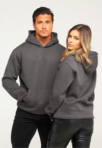 Graue Kapuzenpullover mit Kängurutaschen; weicher Stoff, oversized Schnitt. Das Modell posiert Rücken an Rücken und trägt schwarze Hosen.