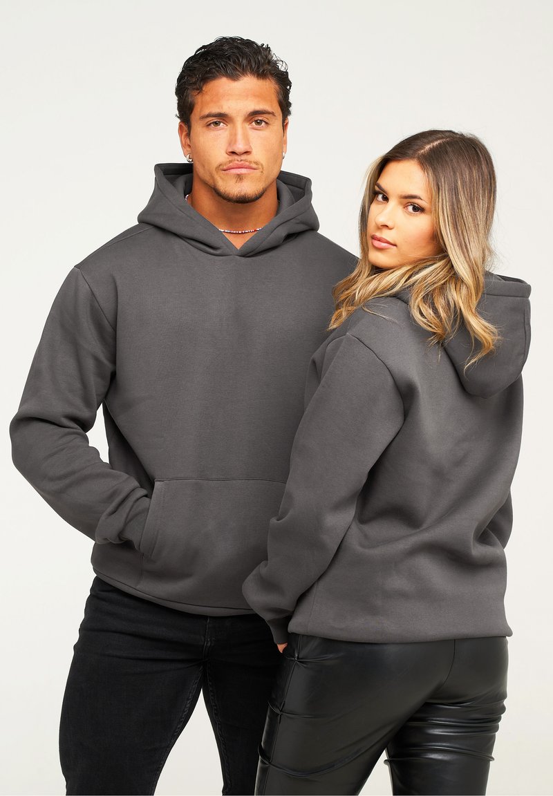 Graue Kapuzenpullover mit Kängurutaschen; weicher Stoff, oversized Schnitt. Das Modell posiert Rücken an Rücken und trägt schwarze Hosen.