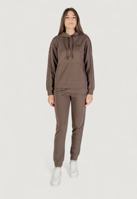 Joven mujer de pie en un chándal marrón de EA7 con sudadera con capucha y pantalones a juego, usando zapatillas blancas sobre un fondo neutro.