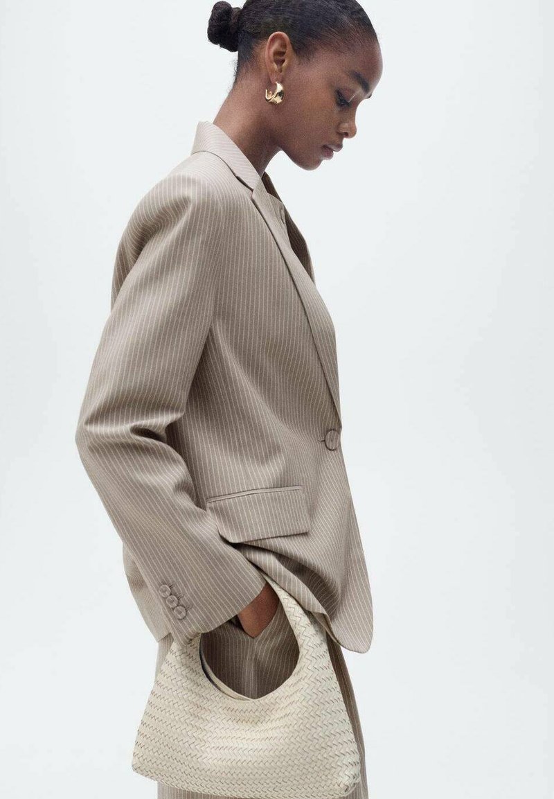 Blazer beige chiaro a righe sottili con una vestibilità sartoriale, abbinato a una borsa intrecciata testurizzata. I capelli del modello sono acconciati in uno chignon elegante.