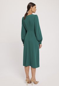 Turkusowa sukienka midi z długimi puff sleeves, zamkiem błyskawicznym z tyłu, subtelnymi plisami i gładką fakturą, zestawiona z cielistymi szpilkami.