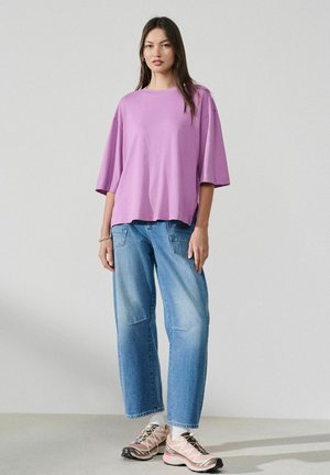 Woman standing wearing a loose lavender t-shirt, light blue wide-leg jeans, white socks, and pink and beige sneakers on a light background.