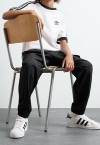 Vit t-shirt med svarta detaljer och tre ränder, svarta avsmalnande byxor och vita sneakers med svarta ränder, sittande på en trästol.