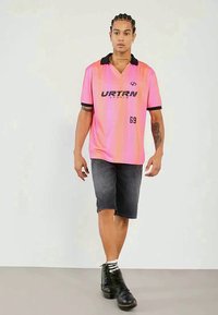 Polo majica s kratkimi rokavi v rožnatem gradientu z črnim ovratnikom in logotipom, v kombinaciji s temno denim kratkimi hlačami in črnimi čevlji. Model stoji v nevtralni ozadju.