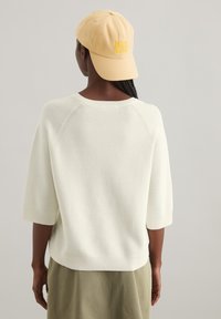 Pull en tricot de couleur crème avec des manches trois-quarts et un col rond, porté avec une casquette brodée jaune clair. Texture lisse.