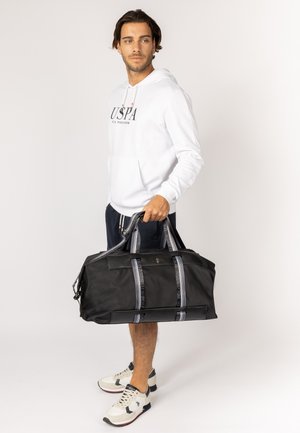 Bolsa de deporte negra con correas de red grises, cierre de cremallera y bolsillo frontal. Textura suave, forma rectangular y detalle destacado del logo de la marca.