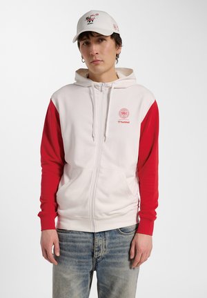 Młody mężczyzna w białej i czerwonej rozpinanej bluzie z kapturem z logo DBU i hummel, biała czapka z grafiką oraz wyblakłe niebieskie dżinsy, stoi na białym tle.