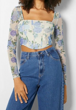 Mujer con un top corsé floral crema con mangas largas transparentes y vaqueros azules de talle alto, mano apoyada en el muslo.