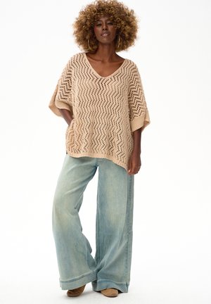 Kobieta z kręconymi włosami, ubrana w beżowy dzianinowy top z dekoltem w serek, jasnoniebieskie szerokie spodnie dżinsowe i beżowe buty, stojąca z jedną ręką w kieszeni.