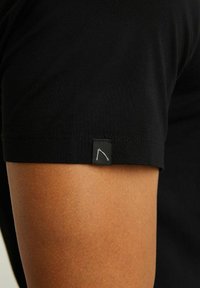 Gros plan sur le bras d'une personne portant un t-shirt noir à manches courtes avec une petite étiquette noire présentant un design minimaliste blanc près de l'ourlet de la manche.