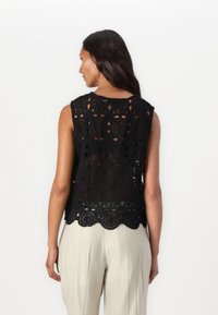 Svart blonde topp med blomster- og geometriske mønstre, ermeløs design, med bølget kant, sett bakfra med nøytrale fargede bukser.