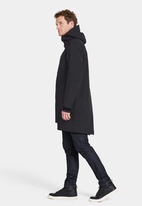 Sort vandtæt parka-jakke med hætte, der har lange ærmer, lige snit og en sidelomme, båret over mørke jeans og sorte sneakers.