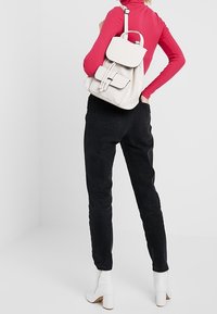 Mochila de cuero blanca con solapa y bolsillo frontal, llevada a la espalda. Modelo con top rojo y pantalones negros de tiro alto, de espaldas.