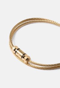 Bracelet en câble torsadé ton or avec un fermoir cylindrique présentant des extrémités texturées sur fond clair.