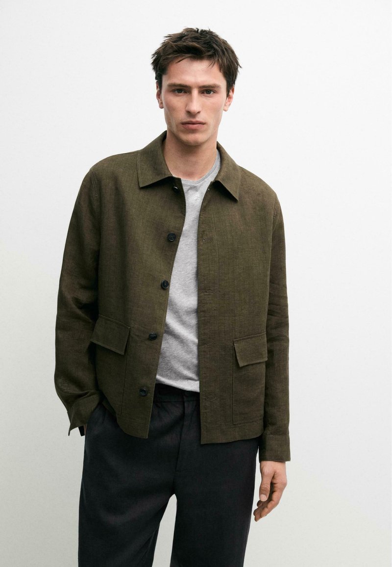 Young man wearing a dark olive buttoned jacket over a light gray shirt and black pants against a plain white background.