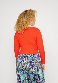 Sweater vermelho com nervuras e mangas longas, combinado com calças estampadas coloridas em tons de azul, roxo e verde. Material tricotado texturizado.