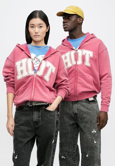 Rosa zip-up-hoodies med texten "HOT" i vitt, kombinerade med grå jeans med små kaninmotiv. Modellen bär en blå skjorta under.