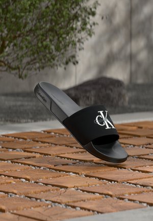 Sandale mule noire Calvin Klein avec logo CK blanc, posée sur un sol en carreaux de bois avec de petits cailloux entre les carreaux, en extérieur.