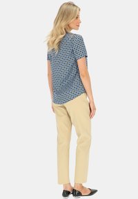 Niebieska bluzka krótki rękaw w geometryczny wzór, zestawiona z beżowymi dopasowanymi spodniami i czarnymi płaskimi butami, widok z tyłu.
