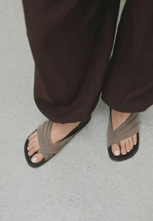 Brązowe płaskie sandały z krzyżującymi się materiałowymi paskami, gładką fakturą i trwałą podeszwą. Palce u stóp mają zadbany pedicure z jasnym lakierem.