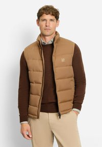 Braune gesteppte Steppweste mit hohem Kragen, Frontreißverschluss und Logo-Patch; getragen über einem dunkelbraunen Pullover; neutralfarbene Hose.