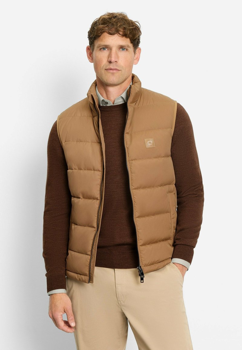 Braune gesteppte Steppweste mit hohem Kragen, Frontreißverschluss und Logo-Patch; getragen über einem dunkelbraunen Pullover; neutralfarbene Hose.