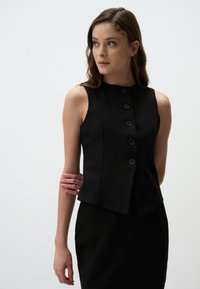 Gilet noir sans manches avec un col rond, doté de six boutons sur le devant, coupe ajustée et tissu lisse, associé à une jupe assortie.