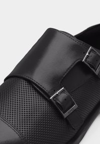 Czarny skórzany but wizytowy z perforowaną teksturą na nosku, podwójnym paskiem typu monk z srebrnymi klamrami oraz gładkim, polerowanym wykończeniem.