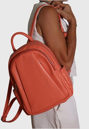 Personne portant un haut blanc sans manches et un pantalon, portant un sac à dos en cuir corail avec deux fermetures éclair et des bretelles ajustables sur une épaule.