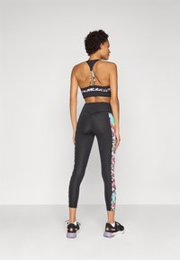Crni sportski crop top s šarenim cvjetnim detaljima i dizajnom s otvorenim leđima, u kombinaciji s crnim tajicama koje imaju cvjetni bočni panel i mrežasti detalj.