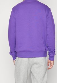 Lila sweatshirt med långa ärmar, ribbade manschetter och nederkant, tillverkad av mjukt material, bärs över ljusgrå sweatpants.