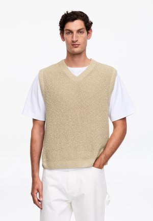 Mężczyzna w beżowym dzianinowym bezrękawniku na białej koszulce i białych spodniach, stojący z jedną ręką w kieszeni na białym tle.