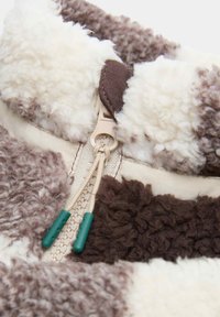 Tela de forro marrón y crema con una superficie texturada y esponjosa. Presenta un zipper con cordones crema y acentos verdes en los extremos.