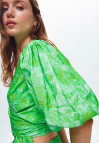 Groene, gestructureerde stoffen top met pofmouwen en een gestrikte taille, met een subtiel bloemmotief doorheen.