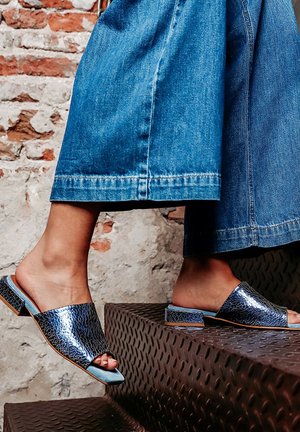 Blue and silver leather slide sandals with a textured finish, open toe design, and low block heel, paired with wide-leg denim.