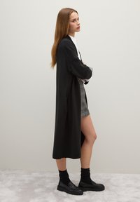 Lungo cardigan nero in un materiale morbido, con maniche ampie e spacchi laterali, indossato sopra una gonna stampata e calzature nere con calzini.