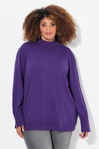 Kvinde med krøllet hår iført en løs lilla sweater og grå bukser, stående mod en ensfarvet hvid baggrund.