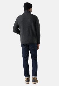 Veste en tricot grise avec col haut, assortie à un jean bleu foncé. Porte un bonnet noir et des chaussures beige avec des détails gris et marron.