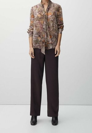 Femme portant un chemisier à manches longues à motifs avec un nœud à l'avant, un pantalon noir à jambes larges et des bottines noires, debout devant un fond uni.