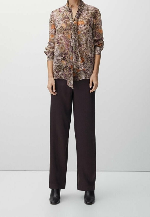 Femme portant un chemisier à manches longues à motifs avec un nœud à l'avant, un pantalon noir à jambes larges et des bottines noires, debout devant un fond uni.