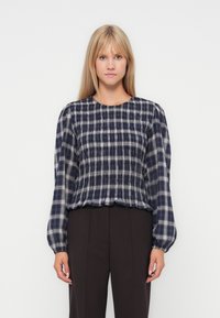 Donna che indossa una blusa a maniche lunghe a scacchi blu navy e bianca con maniche a sbuffo e pantaloni neri, in piedi davanti a uno sfondo bianco uniforme.