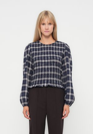 Woman wearing a navy blue and white plaid long-sleeve blouse with puffed sleeves and black pants, standing against a plain white background.