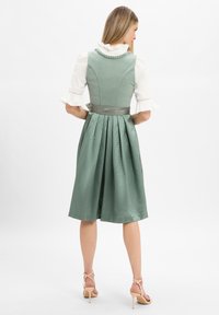 Frau steht mit dem Rücken zur Kamera, trägt ein salbeigrünes plissiertes Kleid mit weißen Rüschensleeves und beigen high-heeled Sandalen.