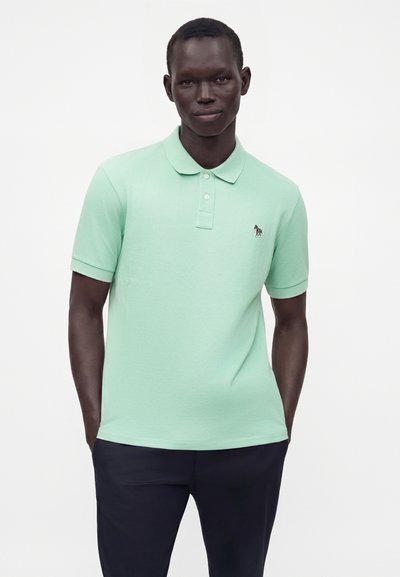 Homme portant un polo vert clair à manches courtes avec un petit logo zèbre brodé, debout les mains dans les poches d'un pantalon foncé.