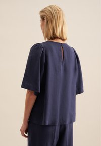 Top in lino blu navy con maniche corte, caratterizzato da un'apertura a chiave e chiusura con bottone sul retro. Vestibilità rilassata e texture morbida.